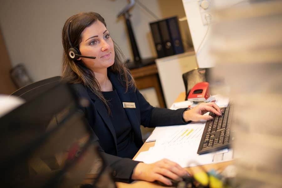 Frau sitzt mit Headset beim Schreibtisch und schaut in den Computer.