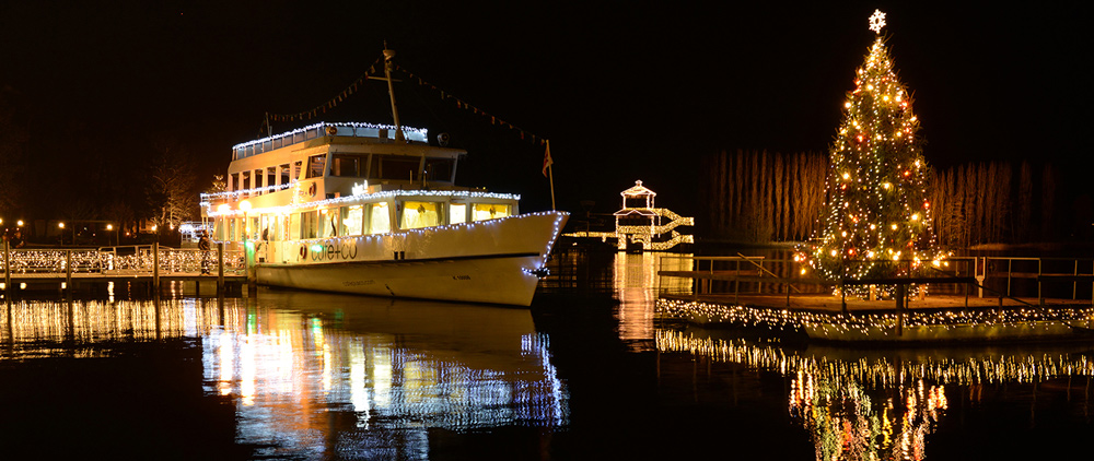 Schiffsanlegestelle in Poertschach bei Nacht. Das Schiff und der Christbaum am Wasser sind weihnachtlich beleuchtet.