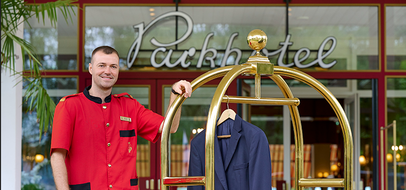 Lohndiener in roter Uniform steht mit einem Gepäckswagen vor dem Parkhotel Pörtschach Eingang