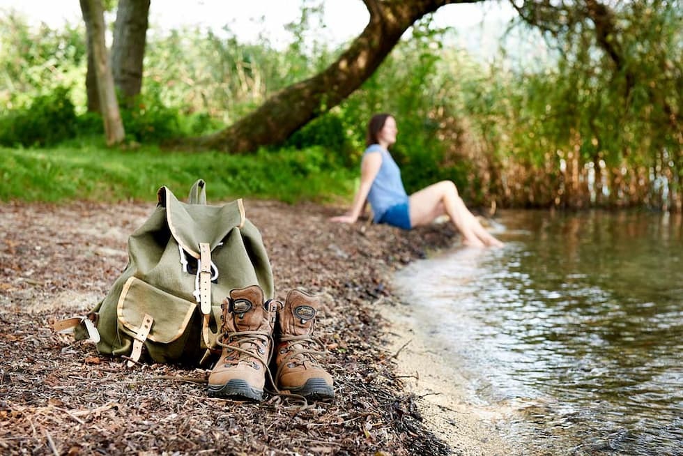 Rucksack mit Wanderschuhe im Vordergrund und Frau sitzt am Seeufer mit den Fuessen im Wasser.